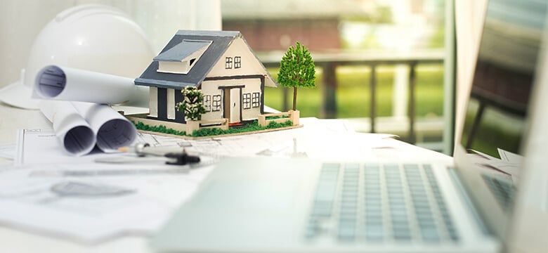 Maqueta de una casa sobre planos, junto a un casco de obra y una laptop
